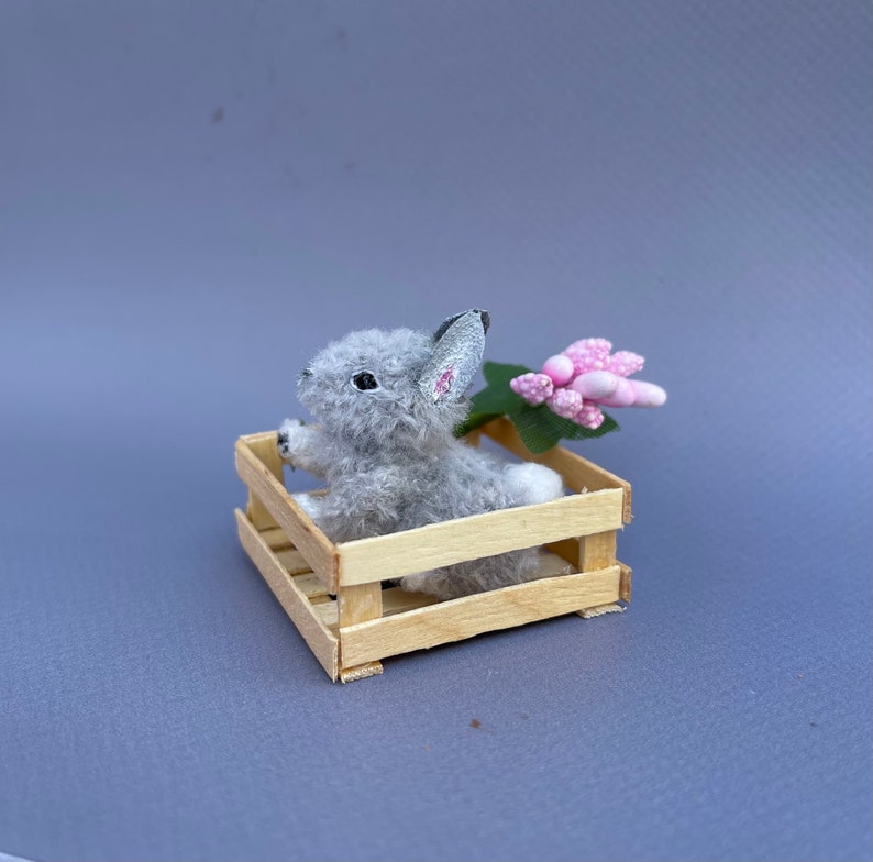 May include: A small gray bunny rabbit sits in a wooden crate with a pink flower.