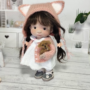 May include: A handmade doll wearing a pink knitted cat ear hood, matching cardigan, and a white dress. The doll holds a small brown teddy bear and wears gray shoes with flower details. The doll has dark hair and is posed in front of a miniature dollhouse.