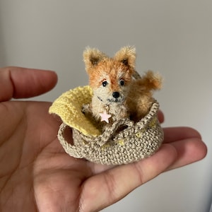 May include: A miniature, handcrafted dog figurine with brown and tan fur, resting in a small, crocheted beige bag with a yellow lining. The dog wears a collar with a pink star charm. The bag is held in a hand.
