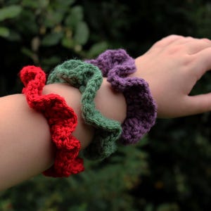 May include: Three crocheted hair scrunchies in red, green, and purple. The scrunchies are worn on a person's wrist.