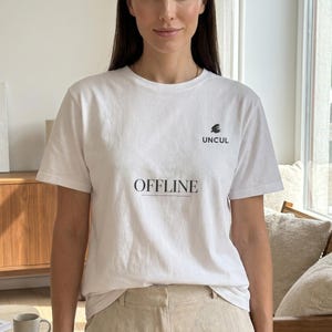 Op de afbeelding: Een wit T-shirt met korte mouwen en een ronde hals, voorzien van een klein zwart grafisch logo met de tekst UNCUL op de linkerborst en het woord OFFLINE in het zwart met een horizontale lijn eronder in het midden van de borst.