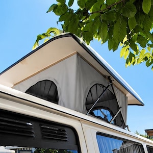 Può includere: Un camper color crema dotato di tetto a soffietto con pareti in tessuto grigio e cornici dei finestrini nere. La carrozzeria presenta una sottile striscia orizzontale arancione e gialla lungo il fianco, con maniglie nere e vetri oscurati.