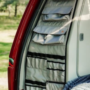 May include: A grey fabric storage organizer mounted on the interior side panel of a red vehicle. The organizer features multiple horizontal pockets with flaps at the top and a series of vertical elastic loops and zippered compartments at the bottom.