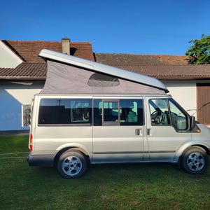 May include: A silver camper van with a pop-up roof tent featuring grey fabric sides and a small window. The vehicle has tinted side windows, black trim, and silver alloy wheels, shown from a side profile view against a clear blue sky.
