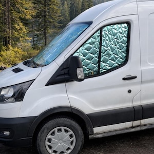 Op de afbeelding: Witte bestelwagen met een zilveren gewatteerde raambekleding geparkeerd in de buitenlucht. De raambekleding heeft een ruitpatroon. De bestelwagen staat op een onverharde weg met bomen en bergen op de achtergrond.