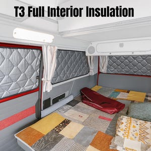 以下が含まれることがあります： 「T3 Full Interior Insulation」の文字が入ったバンの内装。キルティング加工されたグレーの窓カバー、カーテン、様々な色のパッチワークの寝具が特徴です。赤いバッグがベッドの上に置かれ、照明器具が見えます。