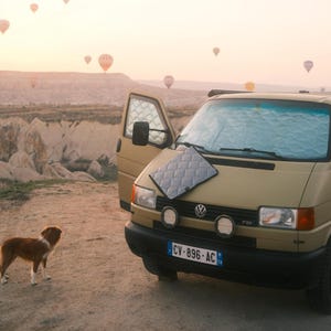 May include: A tan Volkswagen van with a quilted gray thermal window cover resting on the hood and another installed on the windshield. The van features round auxiliary lights, a black bumper, and a French license plate reading CV-896-AC.
