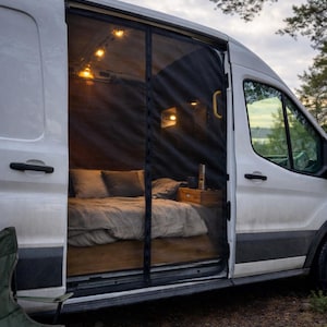May include: A white camper van with the side door open, showcasing a comfortable interior. Inside, a bed with grey bedding and pillows is visible, alongside a small wooden bedside table. String lights illuminate the space, creating a warm atmosphere. A black screen door is in place.