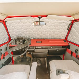 Pode incluir: Vista interior de uma carrinha camper vermelha e cinzenta vintage. O painel de instrumentos, o volante e os bancos são visíveis. As janelas estão cobertas com capas isolantes brancas acolchoadas. O interior da carrinha está bem conservado e limpo.