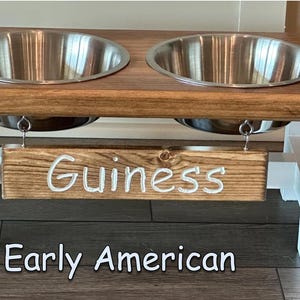 May include: A wooden dog bowl stand with two stainless steel bowls. The stand is painted white and has a wooden sign that says "Guinness" hanging from it. The stand is labeled "Early American".