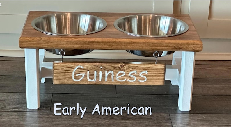 May include: A wooden dog bowl stand with two stainless steel bowls. The stand is painted white and has a wooden sign that says "Guinness" hanging from it. The stand is labeled "Early American".