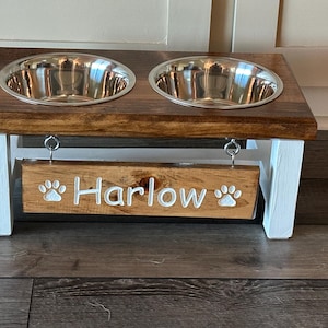 May include: A wooden pet food stand with two stainless steel bowls and a sign that says "Harlow" with paw prints on either side.