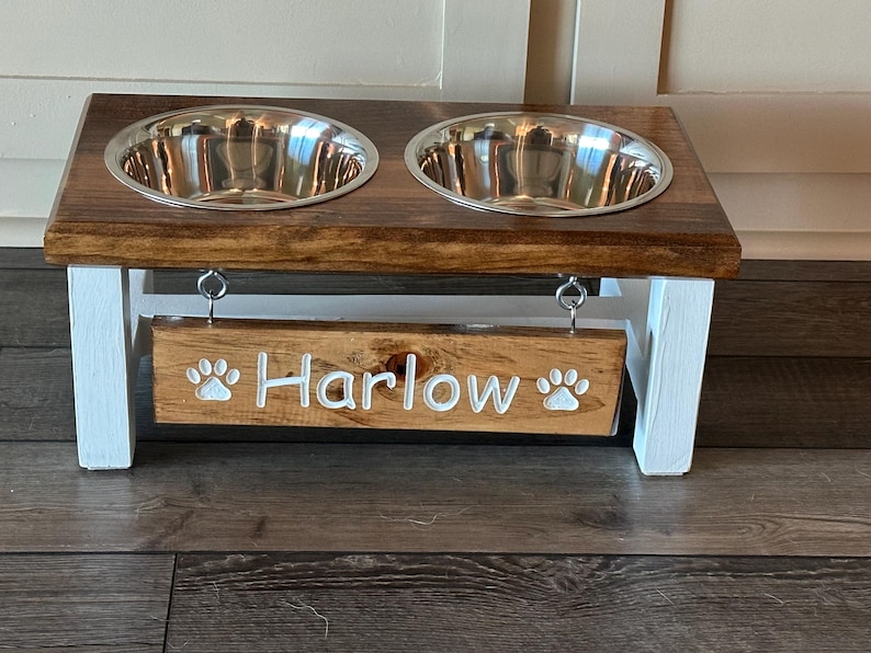 May include: A wooden pet food stand with two stainless steel bowls and a sign that says "Harlow" with paw prints on either side.