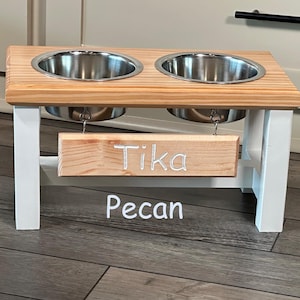 May include: White wooden double dog bowl stand with two stainless steel bowls. The stand has a wooden sign that says "Tika Pecan".