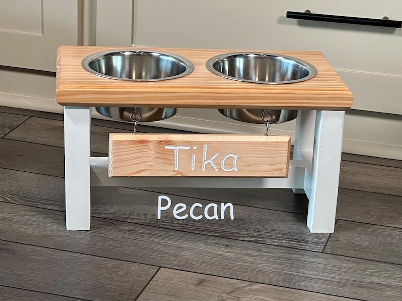 May include: White wooden double dog bowl stand with two stainless steel bowls. The stand has a wooden sign that says "Tika Pecan".