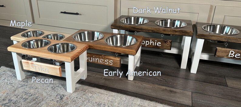 May include: Four elevated dog bowls in different wood finishes: Pecan, Early American, Dark Walnut, and Maple. Each bowl has a personalized name tag: Tiko, Guinness, Sophie, and Belle.