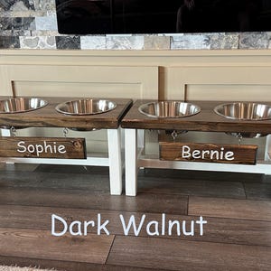 May include: Double elevated dog bowl stand in dark walnut with white legs and stainless steel bowls. The stand has two personalized signs that read "Sophie" and "Bernie".