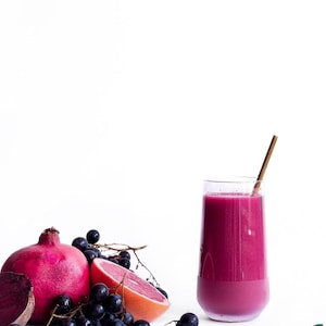 May include: A glass of purple smoothie with a gold-coloured straw, next to a pomegranate, grapes, a slice of grapefruit, and a beet slice. The ingredients are arranged on a white surface. The image has a bright, clean aesthetic, with a focus on fresh produce.