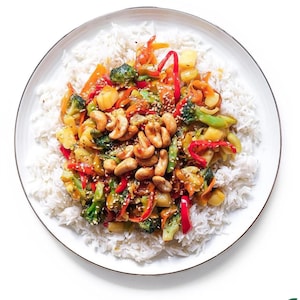 Puede incluir: Un plato de arroz blanco cubierto con un salteado de verduras, anacardos y semillas de sésamo. Las verduras incluyen brócoli, pimientos rojos y zanahorias.