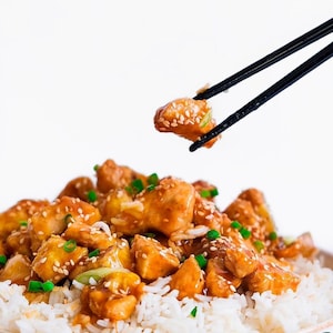 May include: A plate of chicken and rice with a sesame seed and soy sauce glaze. The chicken is being lifted with chopsticks.