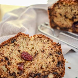 Può includere: Un primo piano di una fetta di banana bread con gocce di cioccolato, mirtilli rossi e noci. Il pane è di colore marrone chiaro e ha una consistenza leggermente friabile.