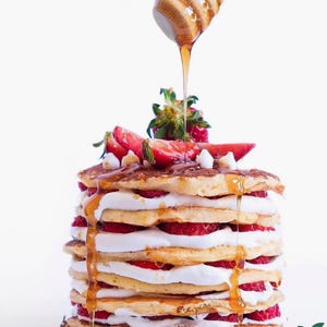 May include: A stack of pancakes layered with strawberries and whipped cream, drizzled with honey. The pancakes are topped with fresh strawberries and a wooden honey dipper. The image is set against a white background.