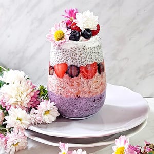 May include: A layered dessert in a clear glass, featuring a purple base, a pink middle layer with strawberries, and a chia seed layer topped with berries and flowers. Pink and white flowers are arranged around the glass on a white plate.