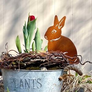 Può includere: Un paletto da giardino in metallo arrugginito a forma di silhouette di coniglio con la scritta "PLANTS" su un vaso di metallo zincato con un nido di ramoscelli e un bocciolo di tulipano rosso.