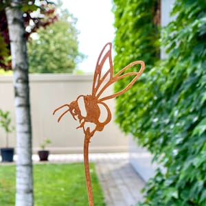 May include: A rusty metal bee silhouette garden stake with a long stem. The bee is facing left and has its wings spread. The stake is placed in a pot of green and red succulents.