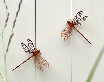 May include: Two metal dragonfly wall sculptures with intricate wing designs. The dragonflies are a rusty orange color and are mounted on a white wall.