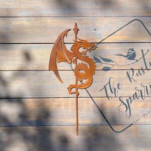 May include: A rusty metal dragon sculpture with wings outstretched, mounted on a metal stake. The sculpture is against a wooden background with a sign that reads "The Rusty Sparrow, The Perfect Gift".