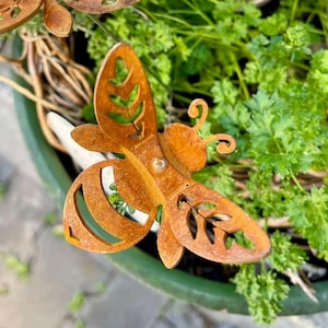 Könnte beinhalten: Zwei rostige Metall-Bienendekorationen mit Blattdetails, platziert in einem grünen Topf mit Kräutern und einem kleinen Keramikkrug. Die Bienen haben ausgeschnittene Flügel und Körperteile, die das Gartenbild verschönern.
