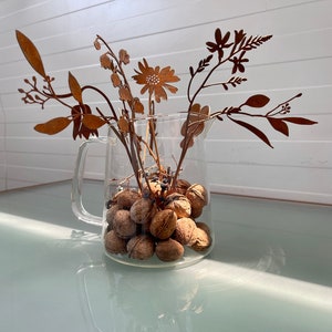 Puede incluir: Una jarra de vidrio transparente llena de nueces y decorada con recortes de metal de flores y hojas.