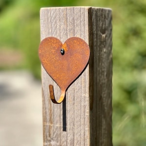 May include: A rusty metal heart-shaped hook attached to a weathered wooden post. The hook is designed to hang items such as keys, towels, or small decorations.