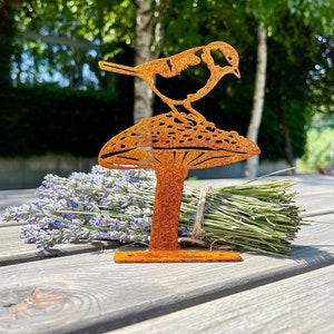 Può includere: Una scultura da giardino in metallo arrugginito di un uccello appollaiato su un fungo. La scultura è su una superficie di legno con un mazzo di fiori di lavanda.