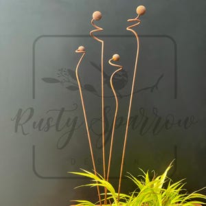 May include: Four rusty metal garden stakes with spiral and ball accents. The stakes are of varying heights and are placed in a pot with green foliage. The background is a dark grey with the text "Rusty Sparrow Designs".