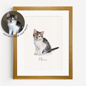 May include: A watercolor painting of a tabby cat named Morris. The cat is sitting with its front paws together and its tail curled behind it. The painting is framed in a light brown wood frame.