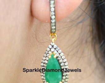 May include: A pair of emerald green gemstone dangle earrings with a pear-shaped stone set in a silver and gold halo of pave diamonds. The earrings are on a model's ear.