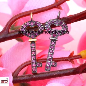 May include: Two key-shaped pendants with a heart-shaped cutout in the head. The keys are adorned with small, round, pink gemstones. One key has a gold-colored top, while the other has a silver-colored top. The background features pink flowers and a brown stem.
