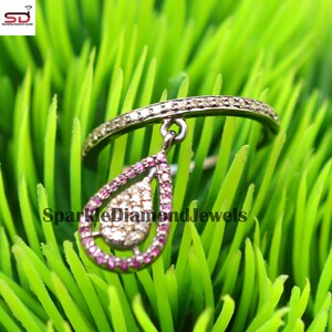 May include: A silver ring with a teardrop-shaped pendant. The pendant is set with small diamonds and has a pink stone border. The ring is on a green background.