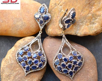 May include: A pair of ornate earrings featuring a teardrop design. The earrings are adorned with numerous oval-cut, deep blue gemstones and surrounded by smaller, clear stones. The metal has a dark, antique finish. The earrings are displayed on a textured, neutral-toned surface.