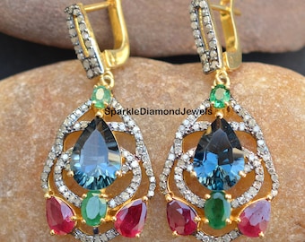 May include: A pair of gold dangle earrings with a teardrop-shaped blue topaz gemstone, surrounded by a silver filigree design and accented with smaller red and green gemstones. The earrings have a hook closure.
