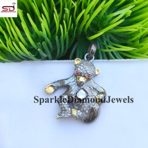 Puede incluir: Un colgante de plata en forma de oso con diamantes negros y ojos de rubí rosa. El oso sostiene un gran diamante transparente en sus patas. El colgante está sobre un fondo blanco.