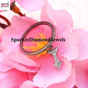 May include: A silver ring with a small diamond-encrusted arrow charm hanging from it. The ring is set against a pink flower background. SparkleDiamondJewels is written in white text on the image.