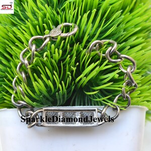 May include: A silver-toned chain bracelet with a rectangular centerpiece and a clasp. The centerpiece is inscribed with the text "SparkleDiamondJewels". The bracelet is set against a backdrop of green foliage.