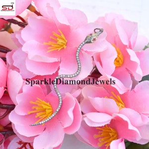 Puede incluir: Un colgante de serpiente de plata con un cuerpo engastado con diamantes y ojos de piedras preciosas rojas. El colgante está fotografiado sobre un fondo de flores rosas.