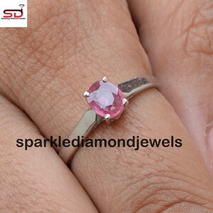 以下が含まれることがあります： ピンクの楕円形の宝石が付いた銀の指輪。指輪は指にはめられています。