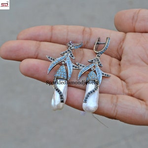 Puede incluir: Un par de pendientes de plata con una perla barroca blanca. Los pendientes tienen un delicado diseño de rama con piedras preciosas negras y azules.