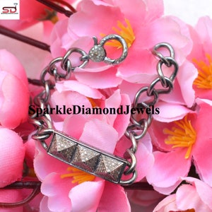 May include: A silver bracelet with a chain link design. The bracelet features a rectangular bar with four pyramid-shaped accents encrusted with small, sparkling stones. The clasp also has small stones. The bracelet is displayed on a bed of pink flowers.