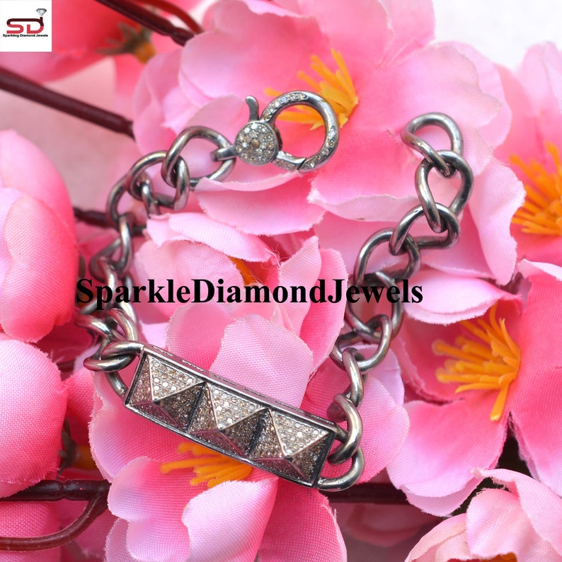 May include: A silver bracelet with a chain link design. The bracelet features a rectangular bar with four pyramid-shaped accents encrusted with small, sparkling stones. The clasp also has small stones. The bracelet is displayed on a bed of pink flowers.